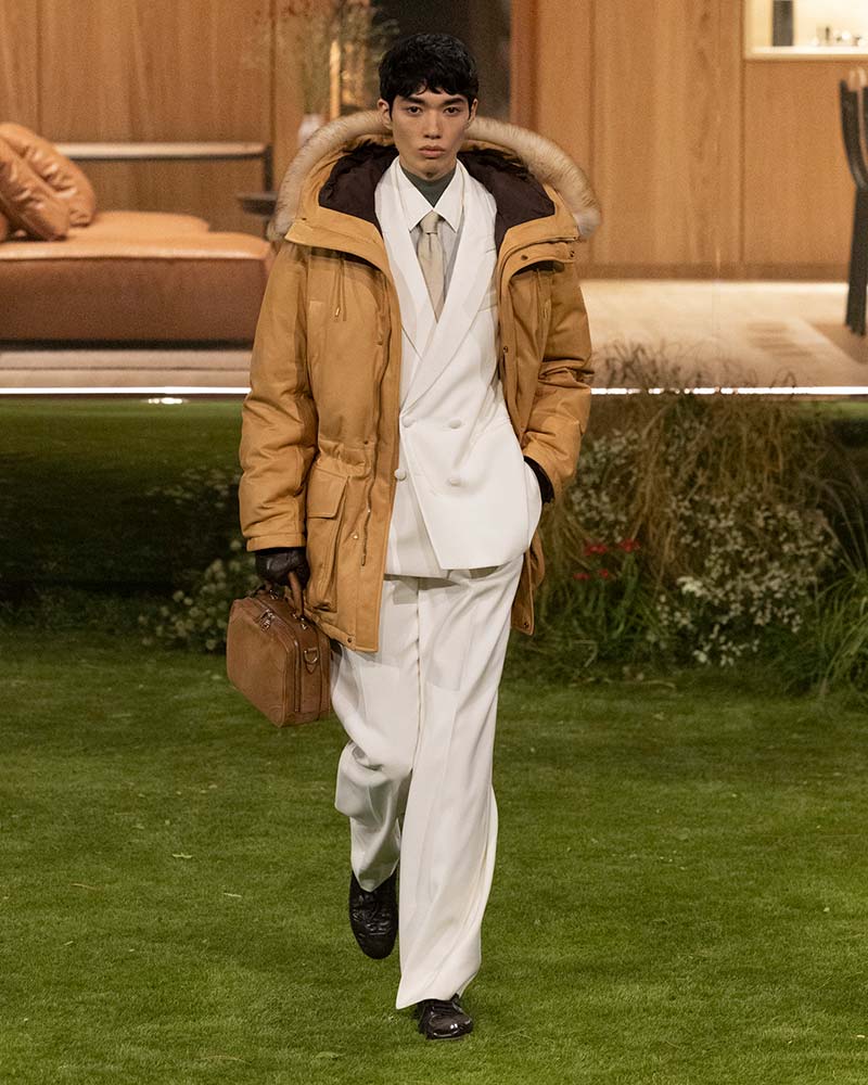 Gebroken wit double-breasted pak van Louis Vuitton met kaneelkleurige parka, olijfkleurig T-shirt, leren tas en zwarte sneakers. Photo courtesy Louis Vuitton